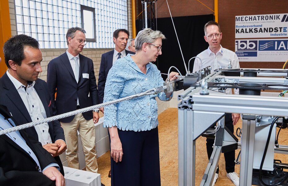 Bundesbauministerin Klara Geywitz betrachtet technische Details des Seilroboters für den Mauerwerksbau